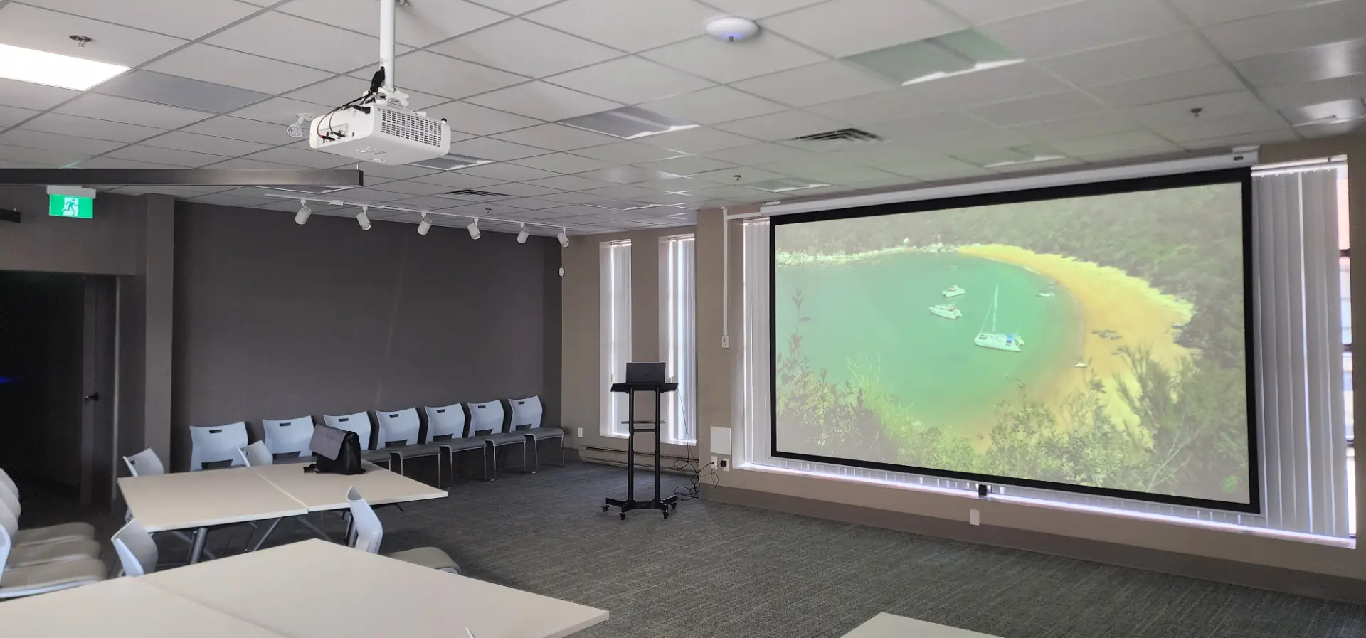 Conference room with a ceiling-mounted projector, drop-down screen showing a tropical bay, and rows of tables and chairs