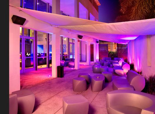 Night-time patio lounge with magenta and purple lighting, sculptural seating and a mounted speaker on a column